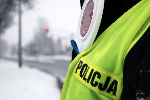 na tle zaśnieżonej drogi widoczna sylwetka człowieka ubranego w żółtą kamizelkę odblaskową z napisem &quot;POLICJA&quot; oraz biało czerwona tarcza do zatrzymywania pojazdów